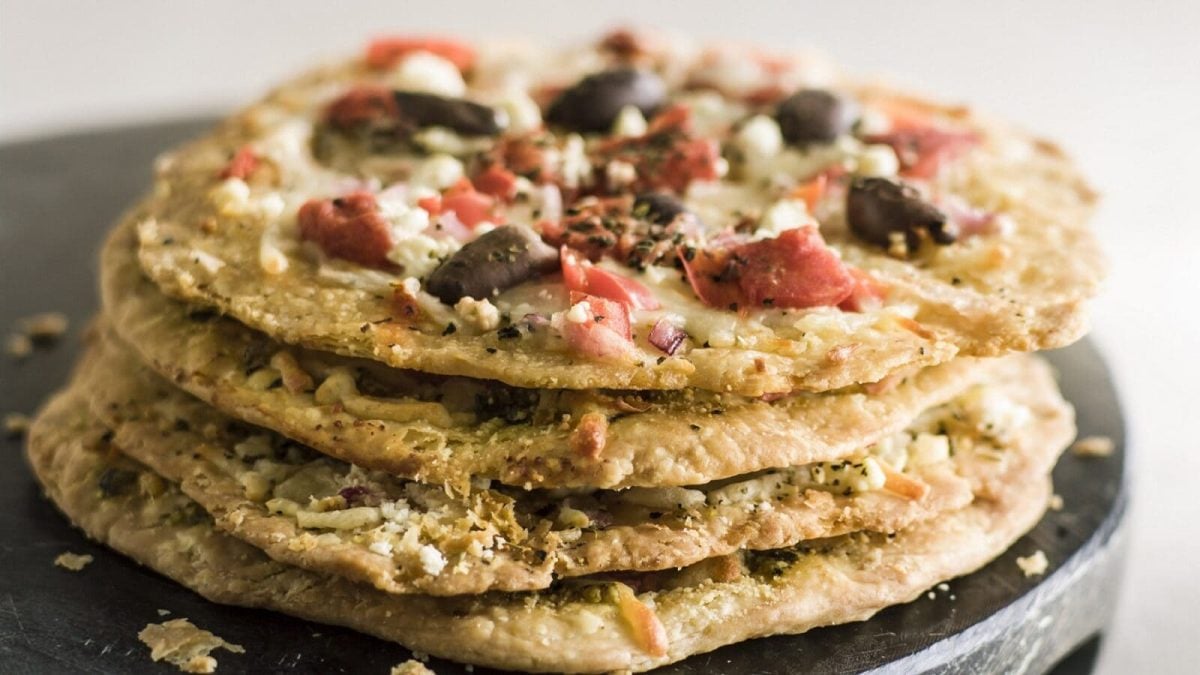 Vier Hüttenkaese-Taler mit Oliven und Tomaten liegen uebereinander auf einem schwarzen Brett auf einem grauen Untergrund.