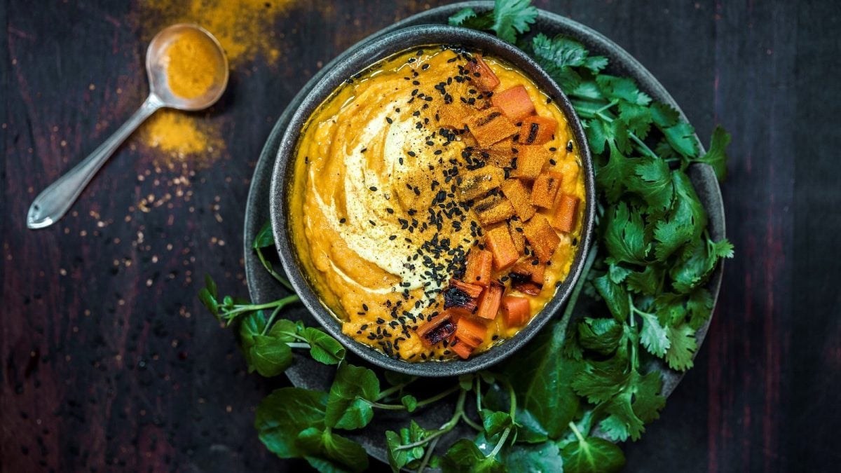 Eine Schüssel mit orangenem Karottendip, garniert mit weißem Crème fraîche und schwarzem Sesam umgeben von frischem Koriander und Kurkuma-Pulver, vor dunklem Hintergrund von Oben.