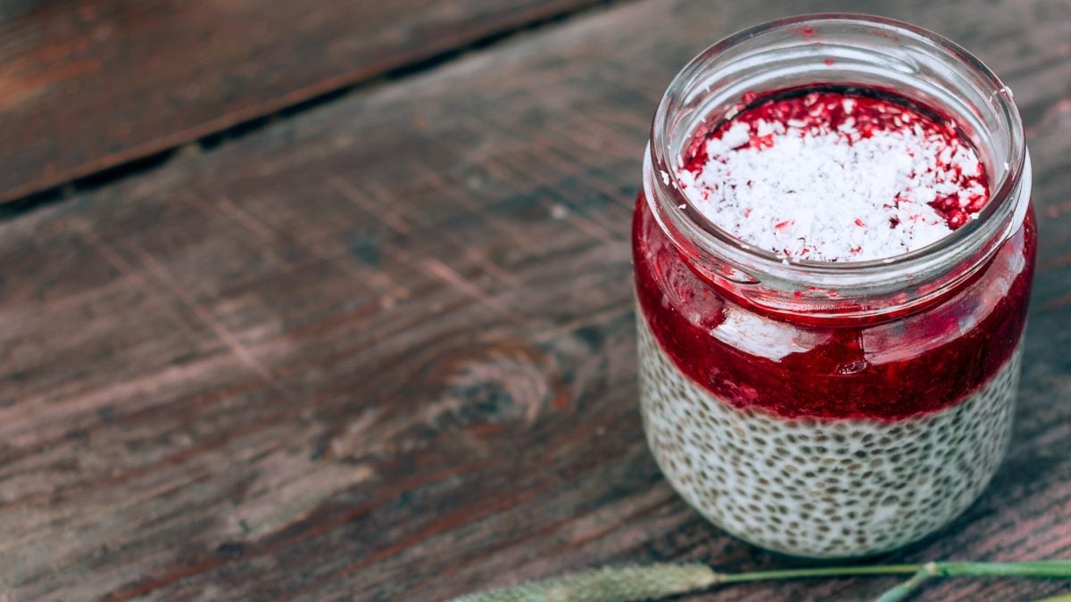 Ein Glas mit Kokos-Chia-Pudding, getoppt mit Himbeerpüree und Kokosraspeln, auf einem Holztisch.