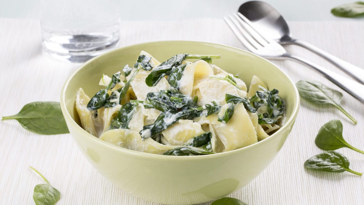 Auf einer weißen Tischplatte steht eine gelbe Schüssel. In dieser Schüssel sind Pappardelle mit Spinat und Ricotta. Auf der Tischplatte liegen noch Besteck, Spinatblätter und darüber hinaus ist noch ein Glas zu sehen.