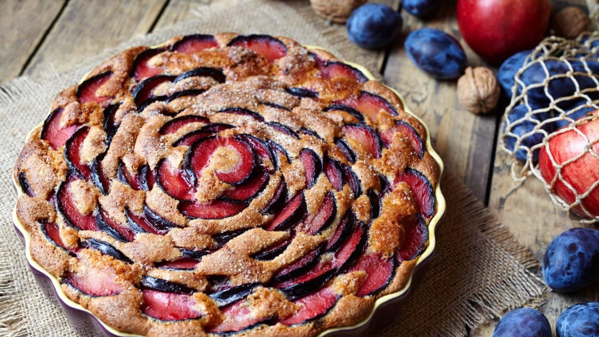 Eine Kuchenform mit Pflaumenkuchen mit Joghurt, daneben liegen Zwetschgen, Äpfel und Nüsse.
