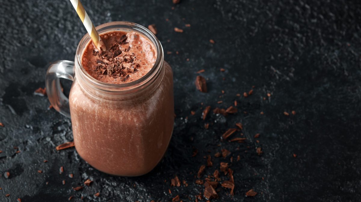 ein Glas Proteinshake mit Schokolade und einem Strohhalb vor dunklem Hintergrund.