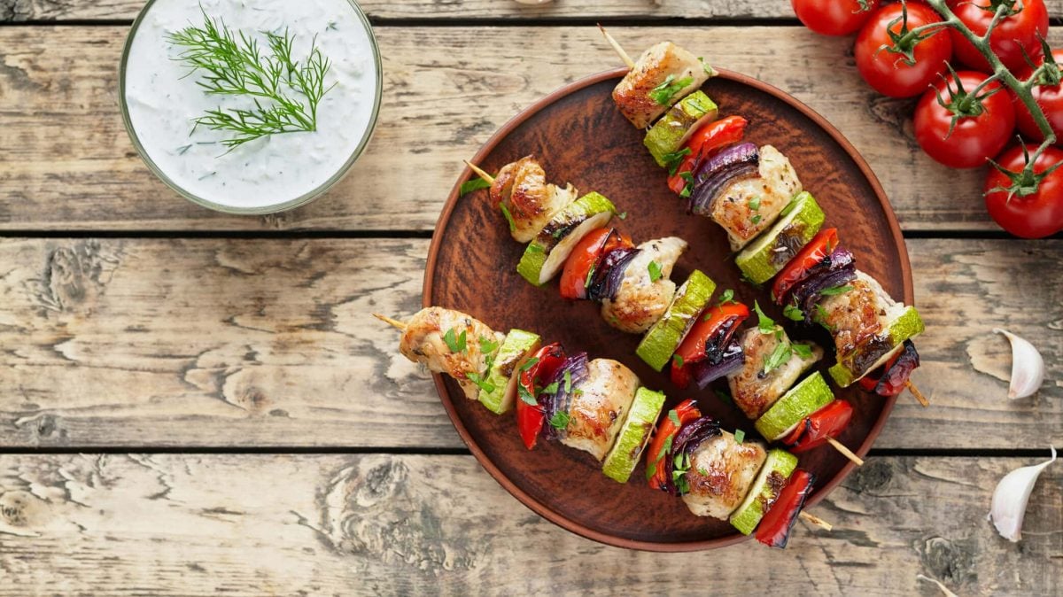 Putenspieße mit Gemüse auf einem dunklen Teller in der Draufsicht. Daneben liegen Strauchtomaten und Knoblauch, außerdem ist eine Schale mit Quark zu sehen.