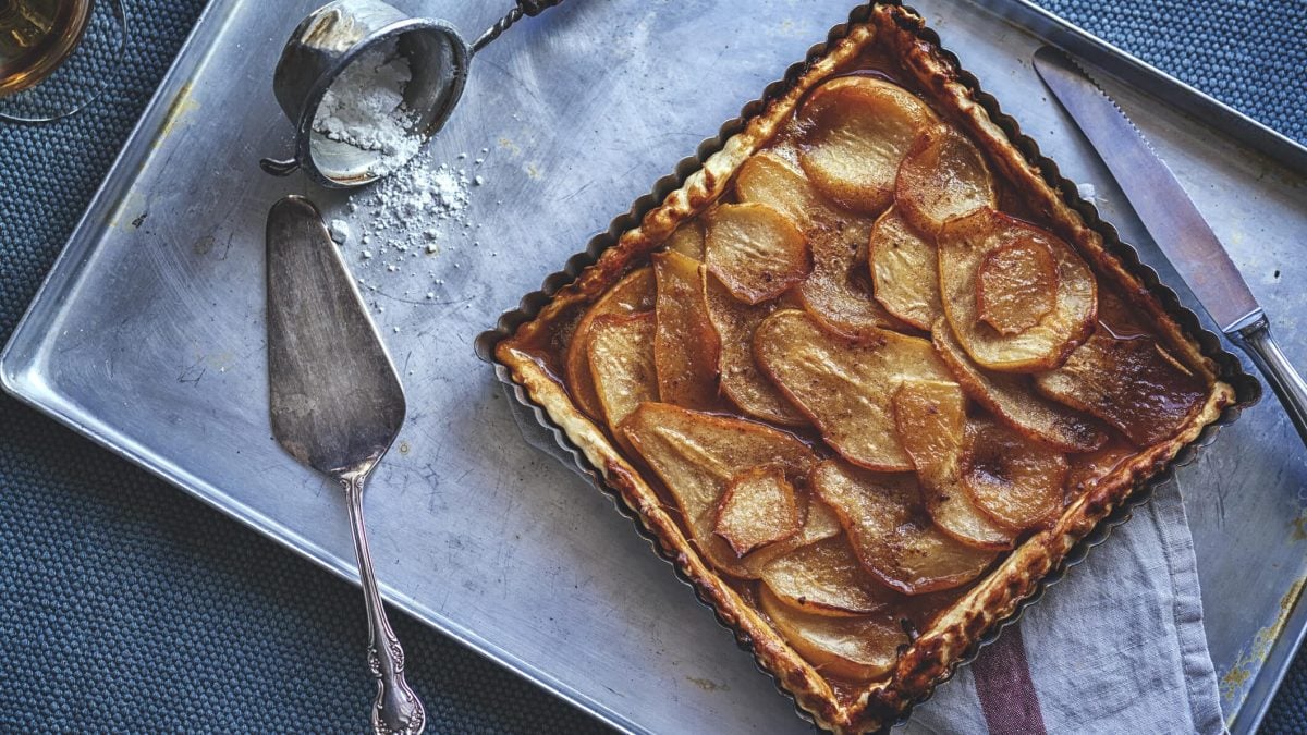 Quark-Birnenkuchen mit Zimt einem Blech, daneben liegen ein Messer, ein Tortenheber und ein Sieb mit Puderzucker.