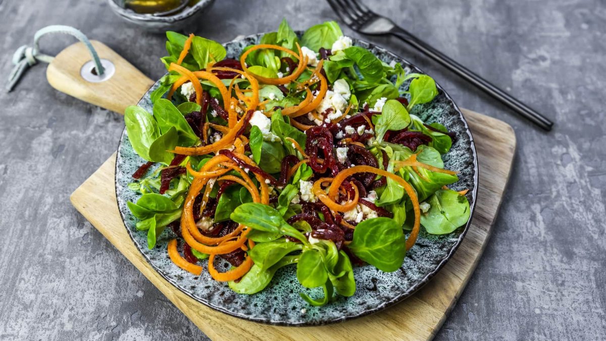 Rote-Bete-Salat mit Feldsalat und Feta auf einem gemusterten Teller. Der Teller steht auf einem Holzbrett. Im Hintergrund liegt eine schwarze Gabel.