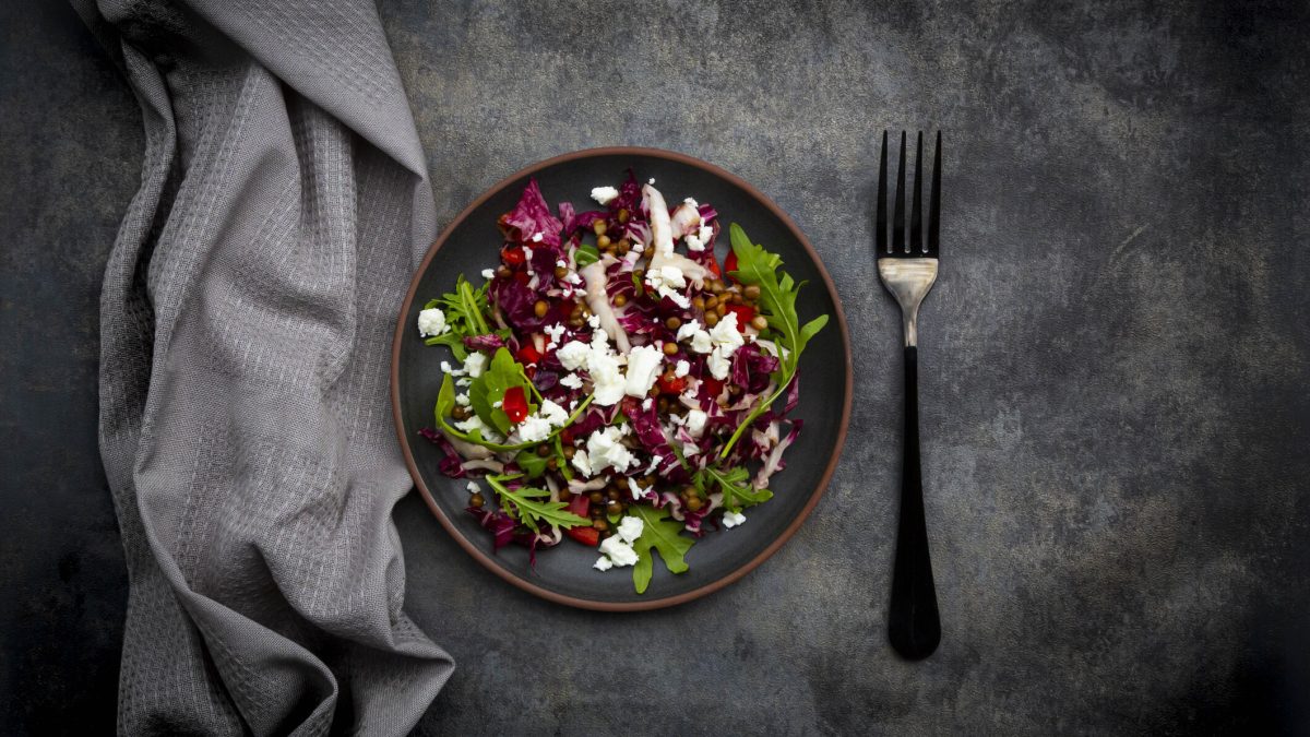Ein Teller mit Rucola-Radicchio-Salat, Feta und Linsen, daneben liegen eine Gabel und eine graue Stoffserviette.