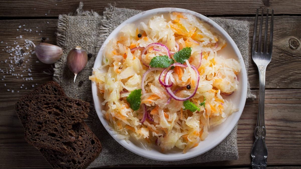 ein heller Teller mit Sauerkrautsalat, daneben liegt eine Gabel, auf der anderen Seite sieht man Knoblauchzehen, Salz und Schwarzbrot. Alles auf Holz.