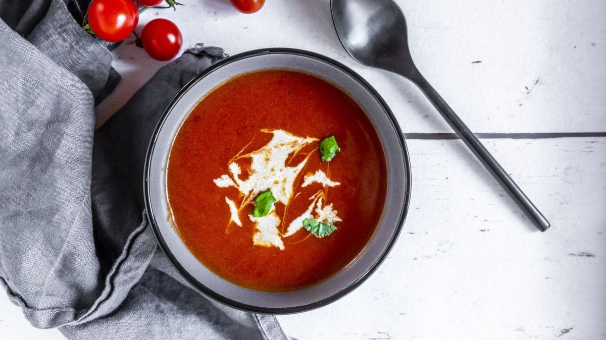 eine Schüssel mit schneller Tomatensuppe, verfeinert mit Basilikum und Joghurt. Daneben liegen Tomaten, ein Geschirrtuch und ein Löffel.