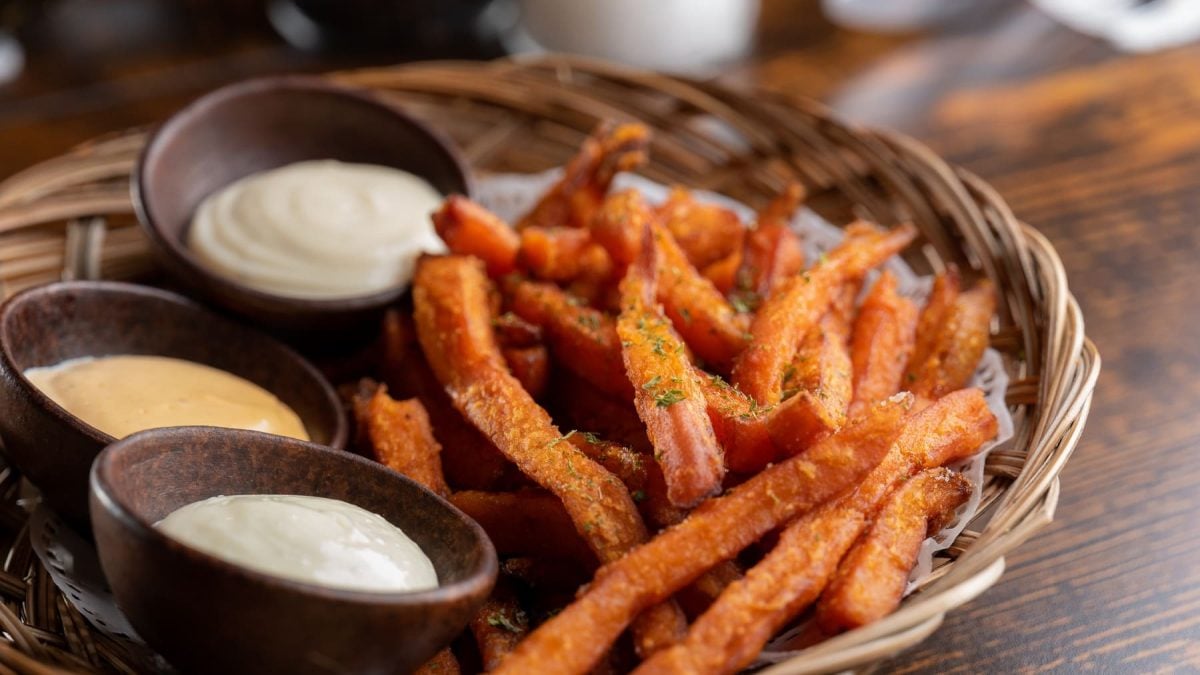 Eine Portion Süßkartoffel-Pommes liegt in einem kleinen Körbchen. Neben den Pommes stehen drei braune Schalen mit verschiedenen Dips. Das Körbchen steht auf einem braunen Holztisch.