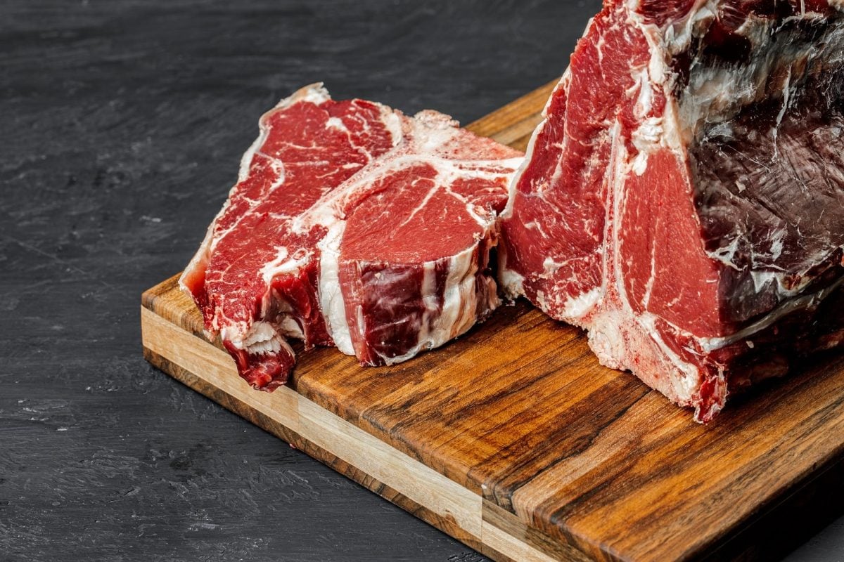 Ein rohes Stück T-Bone-Steak wurde von einem größeren Fleischstück abgeschnitten und liegt auf einem Holzbrett.