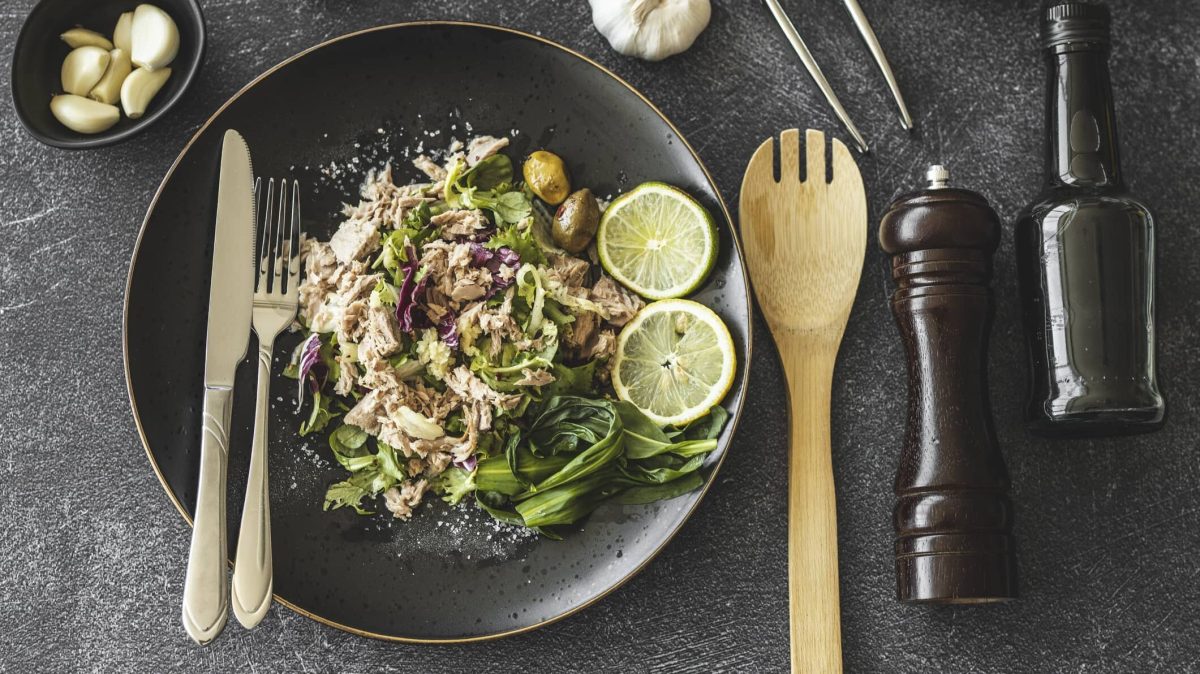 Ein dunkler Teller mit Thunfischsalat mit Oliven, daneben liegt Besteck, außerdem eine Gewürzmühle, eine Ölflasche und ein paar Zutaten.