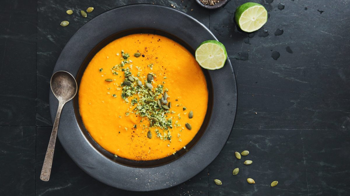 Ein dunkler Teller auf einem dunklen Tisch in der Draufsicht, darin vegane Kürbissuppe, mit Kernen bestreut. Daneben liegen zwei Limettenhälften und ein Löffel.