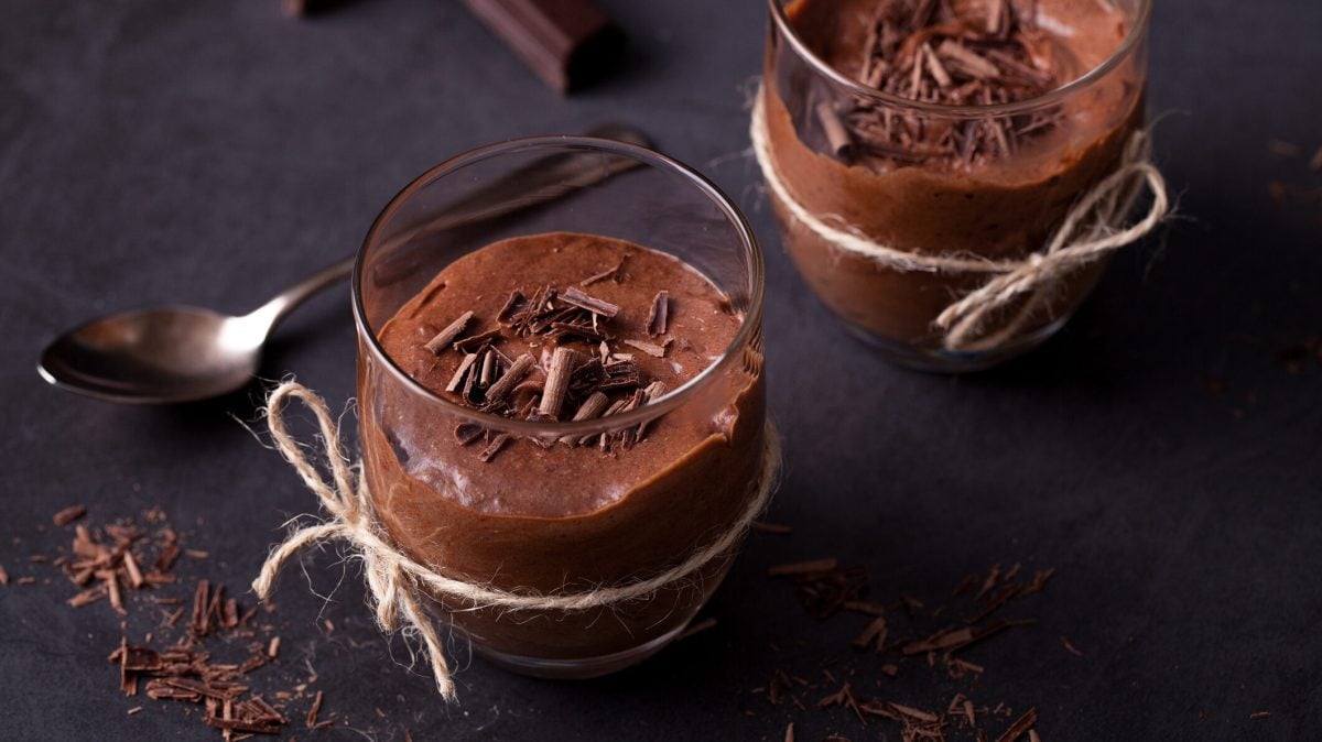 zwei Gläser mit veganer Mousse au Chocolat mit Avocado, dekoriert mit Schokoraspeln, auf einem dunklen Untergrund. Daneben liegen ein Löffel und Schokoraspeln.