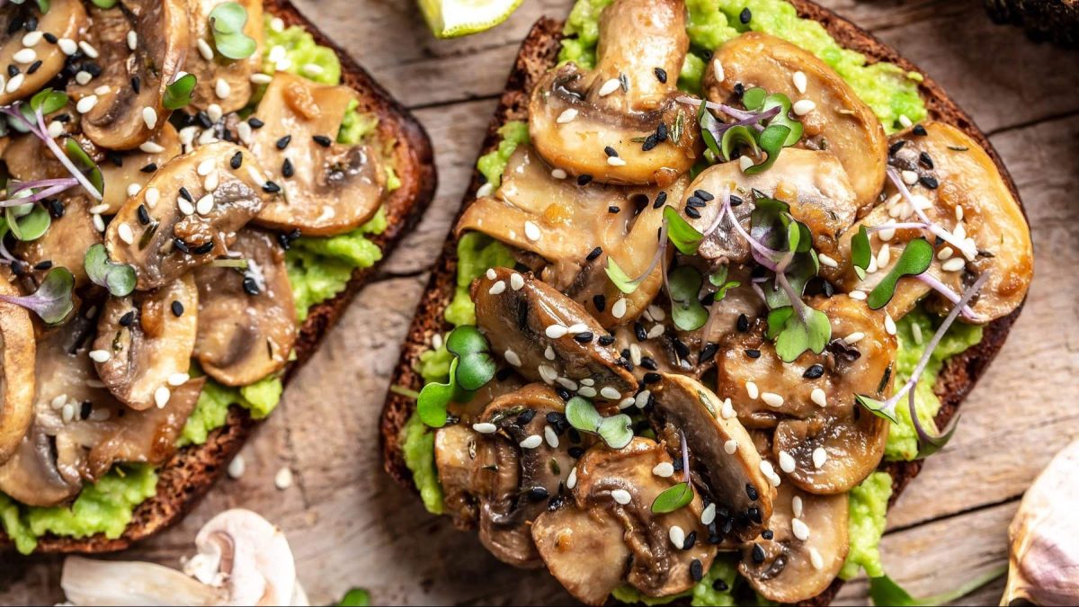Zwei Scheiben Avocado-Toast mit Pilzen auf einem braunen Holztisch. Um die Brote herum liegen Limettenscheiben und Pilze. Das Avocado-Toast ist angerichtet mit Sesam und Sprossen.