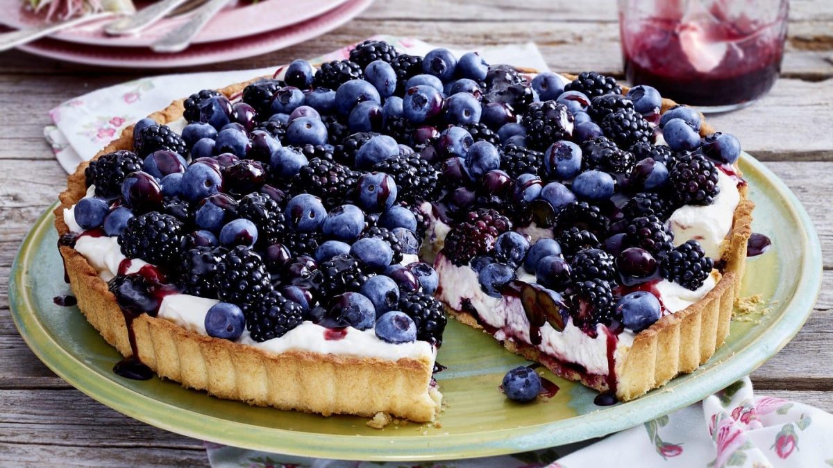 Eine Brombeer-Blaubeer-Tarte auf einem bunten Teller auf einem Holztisch. Um den Teller herum steht ein Glas mit Brombeer-Gelee, Geschirr und Servietten mit Blumendruck.