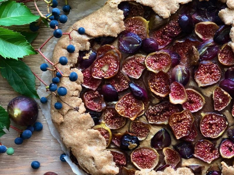Eine Galette mit Feigen und Weintrauben auf einem Holztisch. Um die Galette herum liegen Blätter, Beeren und einige Feigen.