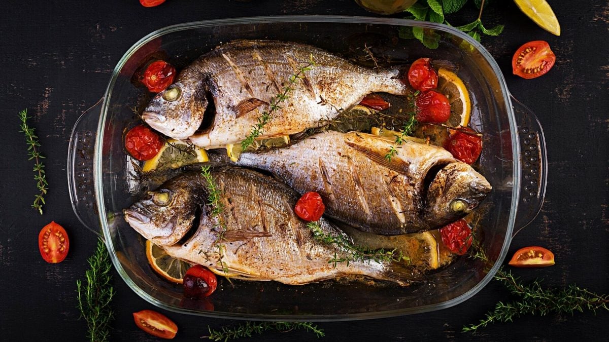 Drei gegrillte Doraden liegen in einer Auflaufform. Neben den Doraden liegen Zitronenscheiben, Thymian und Tomaten. Um die Form herum liegen Tomatenstücken, Zitonenstücken, Thymian. Die gegrillte Dorade ist mit Zitronenscheiben und Kräuterbutter gefüllt.
