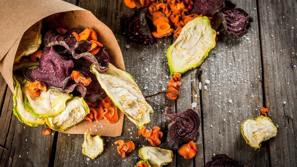 Eine Portion Gemüse-Chips auf einem dunklen Holztisch. Die Chips bestehen aus Zucchini, Möhren und Rote Bete. Auf dem Tisch ist außerdem Salz verteilt.
