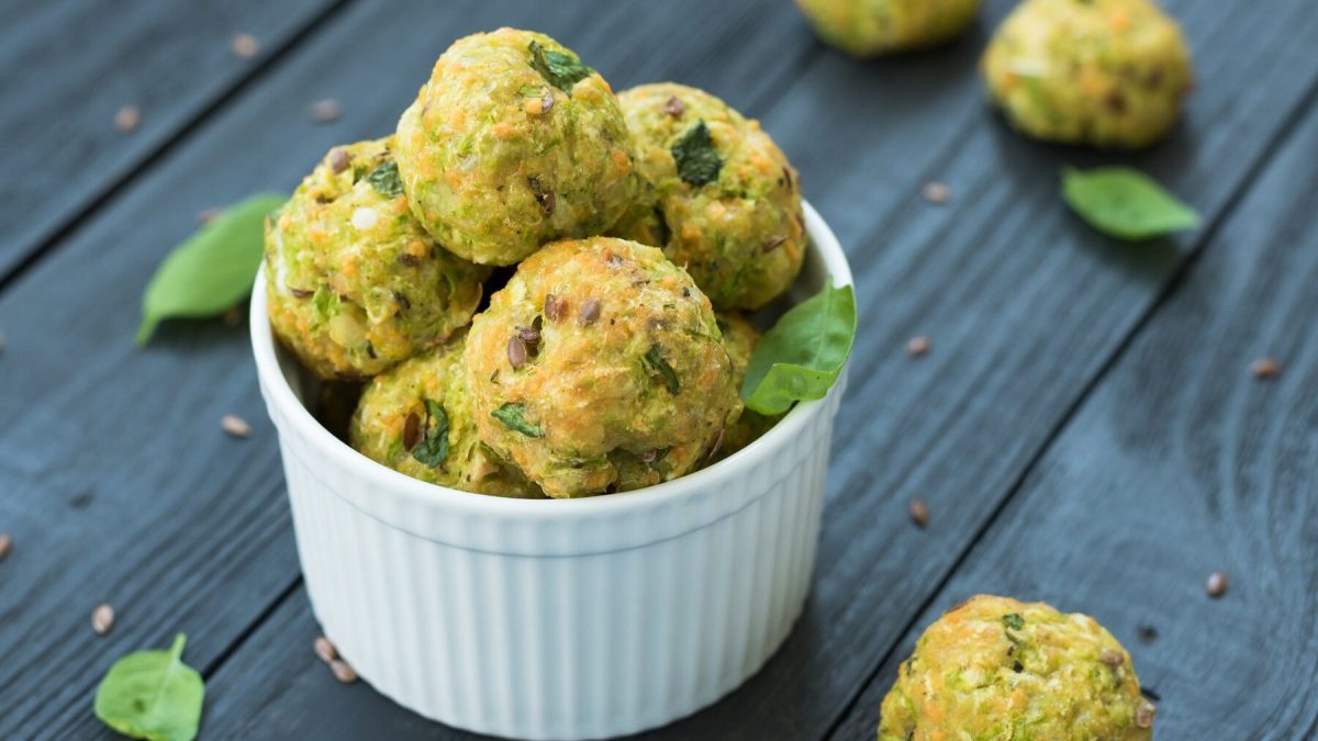 Gemüsebällchen mit Spinat und Parmesan stehen in einer weißen Schüssel auf einem dunklen Holztisch. Die Gemüsebällchen sind angerichtet mit etwas Basilikum und Pinienkernen. Um die Schale herum liegen weitere Gemüsebällchen verteilt.