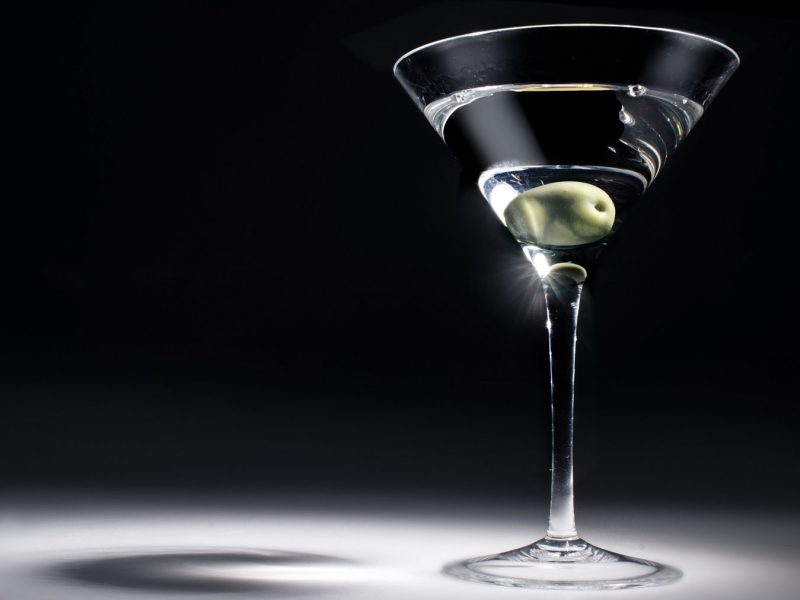James Bonds Martini mit einer Olive in einem Martiniglas. Das Glas wird aus einer Richtung stark beleuchtet, der Hintergrund und Boden sind dunkel.
