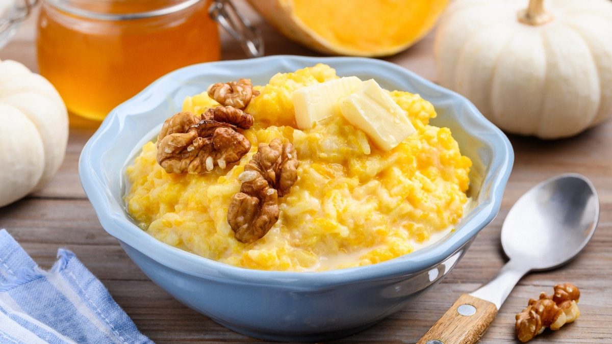 Eine Portion Kürbis-Milchreis mit Nüssen in einer blauen Schale auf einem braunen Tisch. Der Milchreis ist angerichtet mit Walnüssen und Butter. Um die Schüssel herum liegen Walnüsse, ein Löffel, ein Gefäß mit Honig, mehrere Kürbisse und ein blau weißes Geschirrtuch.