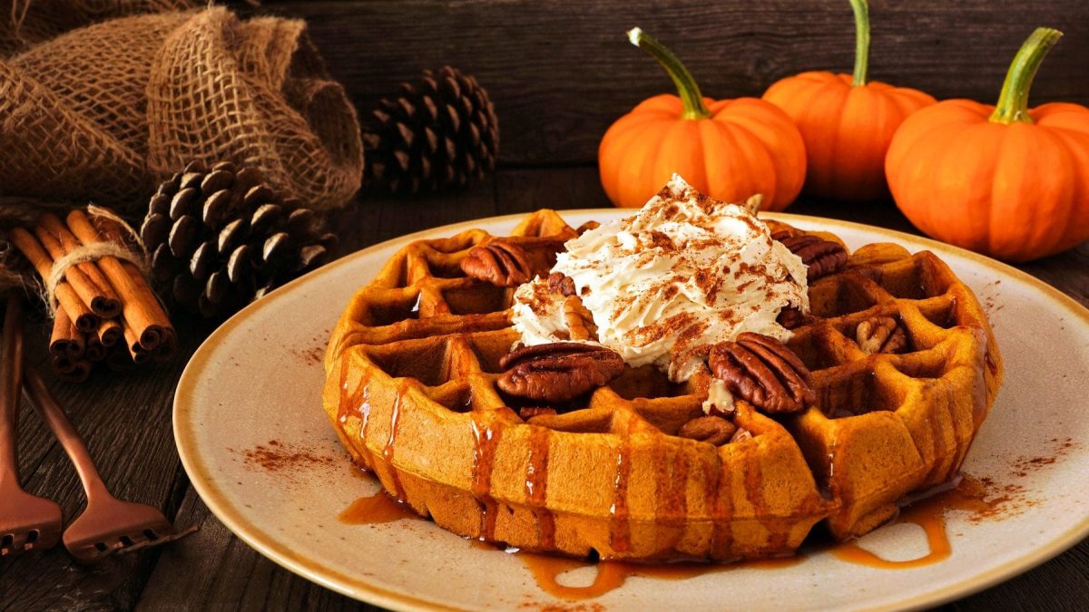 Eine Kürbis-Waffeln mit Pekanüsse, Sahne, Zimt und Ahornsirup liegt auf einem Teller auf einem dunklen Holztisch. Im Hintergrund liegen Kienäpfel, Kürbisse, Zimtstangen, Gabeln und Leinennetze.