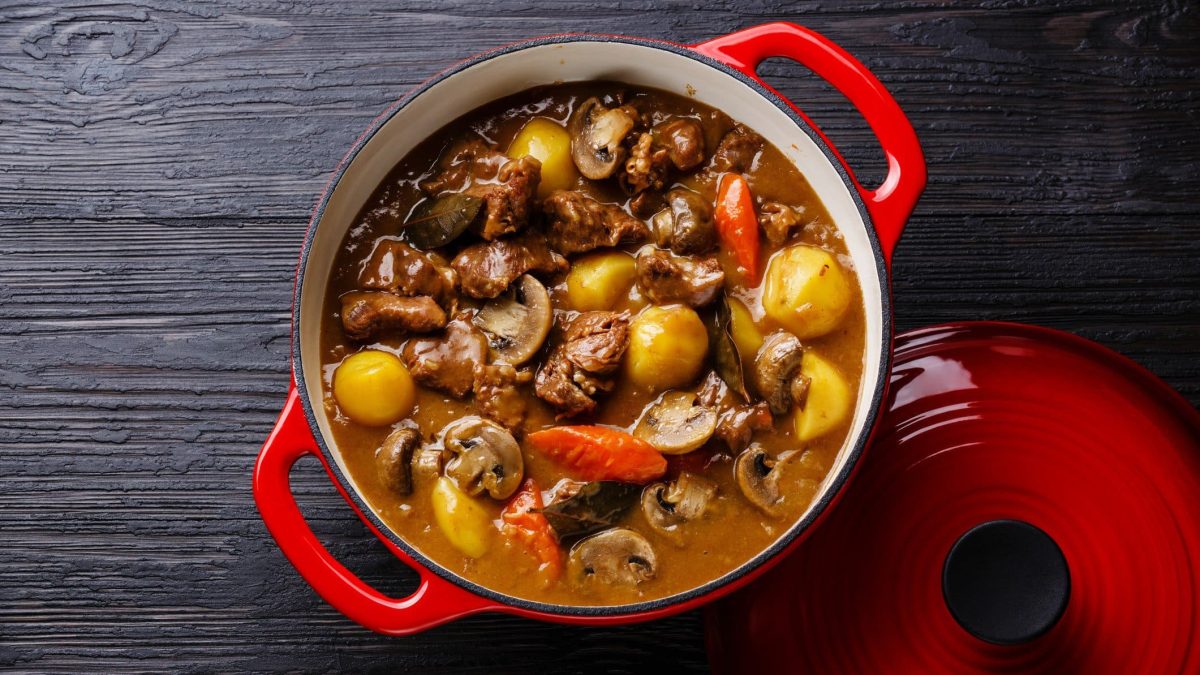 Roter Topf gefüllt mit Pilzgulasch mit Kartoffeln und Gemüse auf dunklem Holzuntergrund.