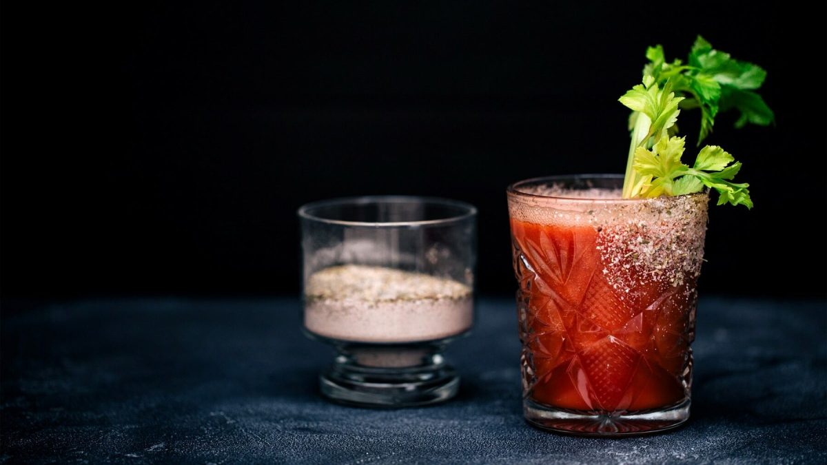 Ein Virgin Bloody Mary in einem Glas mit Selerie. Das Glas ist angerichtet mit Salz und Pfeffer. In einem Glas daneben steht eine Mischung aus Salz und Pfeffer.