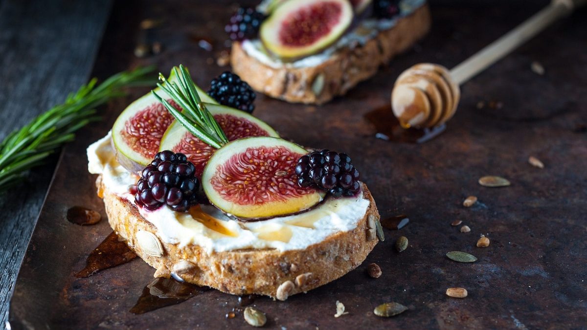 Vollkornbrot mit Feigen, Ricotta und Honig auf einem dunklen Untergrund. Im Hintergrund liegt ein Honiglöffel. Auf dem Untergrund verteilt sind Thymian, Körner und Honig.