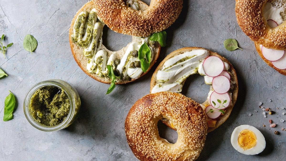Einige halbierte Bagels mit Frischkäse, Pesto und Basilikum liegen auf einem Untergrund aus Stein. Neben den Bagels liegt ein halbiertes, gekochtes Ei, Salz und Pfeffer, Basilikumblätter und ein Gefäß mit grünem Pesto