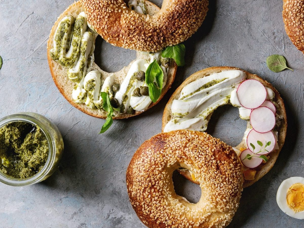 Einige halbierte Bagels mit Frischkäse, Pesto und Basilikum liegen auf einem Untergrund aus Stein. Neben den Bagels liegt ein halbiertes, gekochtes Ei, Salz und Pfeffer, Basilikumblätter und ein Gefäß mit grünem Pesto