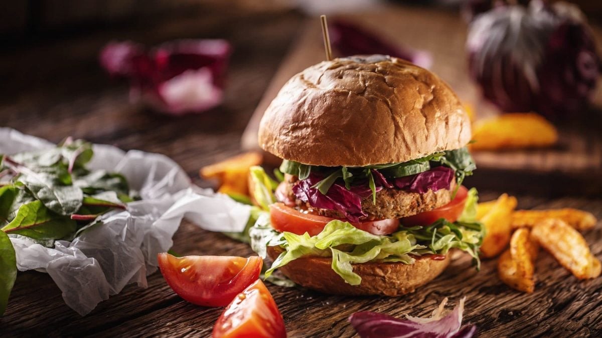 Bellas Gemüse-Burger auf einem Holztisch. Um den Burger herum liegen Pommes, Salat und Tomaten. Der Burger wird mit einem Holzstab zusammen gehalten