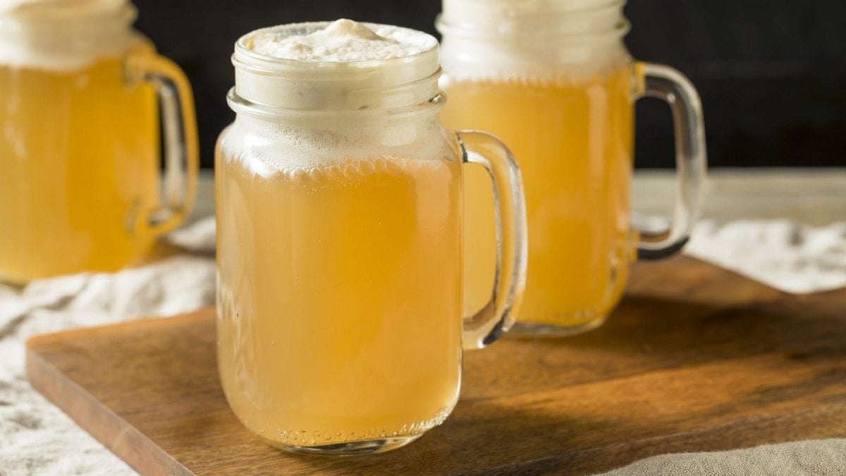 Zwei gläserne Krüge mit Butter-Bier stehen auf einem Holzbrett auf einem Tisch mit einer beigen Tischdecke. Im Hintergrund steht ein weiteres Butter-Bier. Die Biere sind angerichtet mit Sahne.
