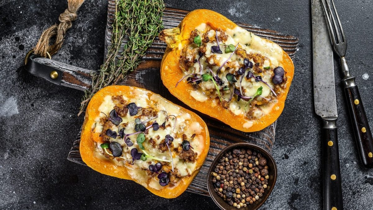 Zwei Hälften Butternutkürbis mit Reis, Hackfleisch unf Pfifferlingen gefüllt, Käse überbacken und frischen Kräutern garniert, auf einem Holzbrett neben Thymian Stengeln und einer Schüssel Pfefferkörner, vor dunklem Hintergrund von oben.
