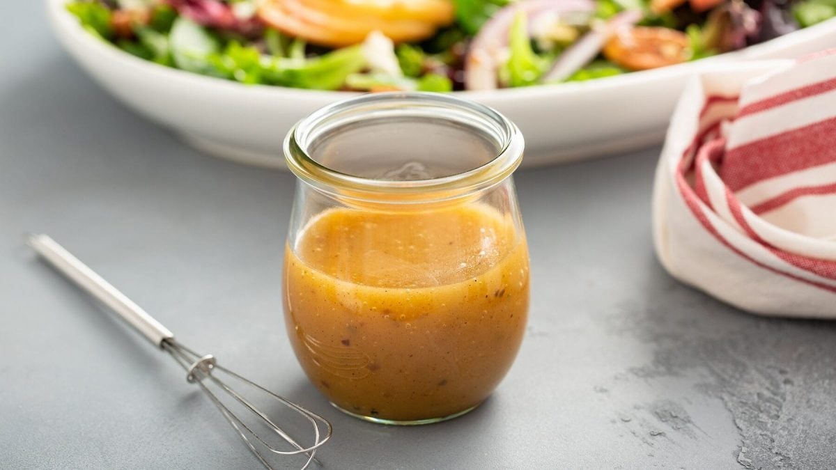 EIn Glas mit Dressing steht auf einem grauen Tisch. Neben dem Glas liegt ein kleiner Rührbesen, im Hintergrund steht eine Schale mit Salat. Am rechten Bildrand befindet sich ein weißes Handtuch mit roten Streifen. Die ultimative Dressingformel.