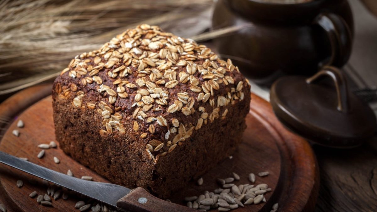 Ein halbes Laib Dreikornbrot in kastiger Form mit Haferflocken und Kürbiskernen bestreut auf einem hölzernen Schneidebrett neben Ehren und einem Tonkrug.