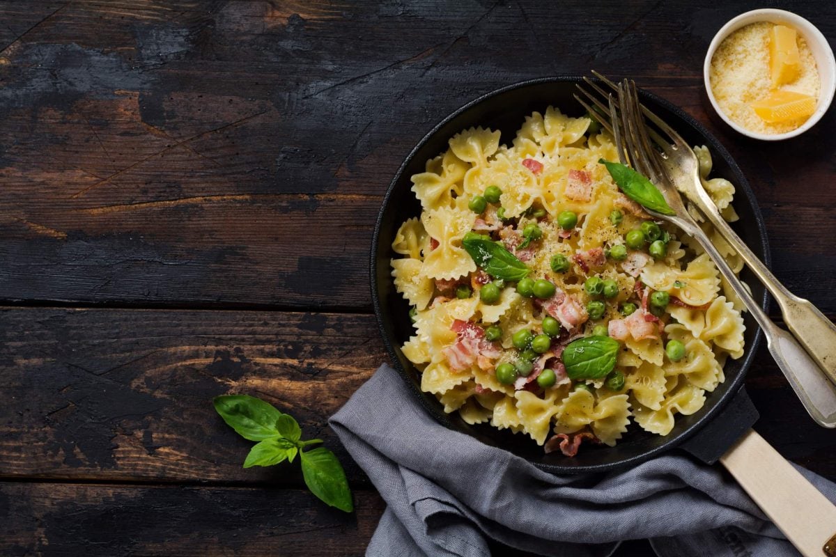 Auf einem Holztisch steht eine Pfanne. In der Pfanne sind Nudel, Schinken, Speck und Basilikum. Daneben steht ein Schälchen mit Parmesan.