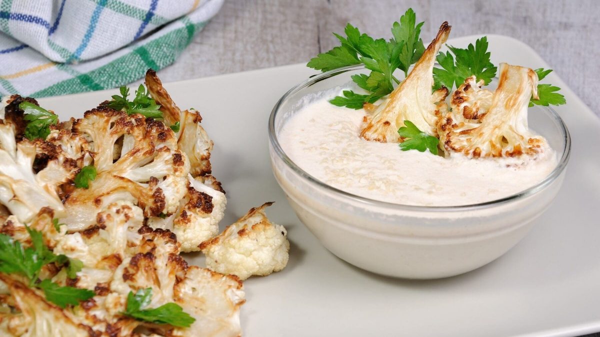 Eine Portion gebackener Blumenkohl mit Tahini-Sauce steht auf einem weißen Tablett auf einem grauen Untergrund. Im Hintergrund liegt ein Geschirrtuch. Der Blumenkohl ist angerichtet mit Petersilie. Die Tahini-Soße befindet sich in einer kleinen Glasschüssel.