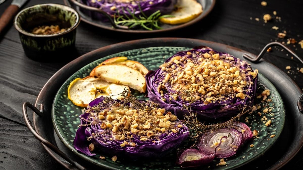Gebackener Rotkohl mit Nüssen und Äpfeln auf einem gemusterten Teller auf einem Tablett. Der Rotkohl ist mit Zwiebelscheiben und gebackenem Apfel angerichtet.
