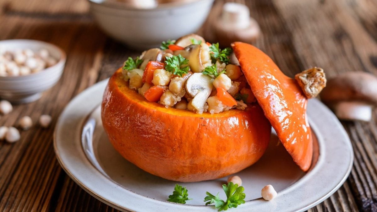 Ein gerösteter Hokkaido-Kürbis gefüllt mit Pilzen, Quinoa und weißen Bohnen auf einem weißen Teller. Der Kürbis ist angerichtet mit Petersilie. Im Hintergrund stehen zwei Schalen mit weißen Bohnen und Pilzen.