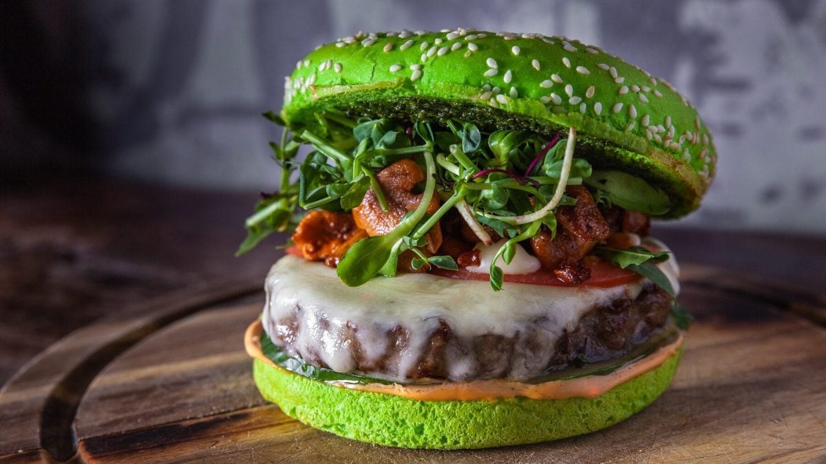 Ein grüner Burger mit Sprossen und Pilzen steht auf einem braunen Holzbrett auf einem hölzernen Untergrund. Der Hintergrund ist grau und gemustert. Auf dem Burgerbrötchen befindet sich Sesam. Der Burger ist außerdem belegt mit einem Burgerpatty, Hackfleisch, einer Tomatenscheibe, Gurke und Soße.