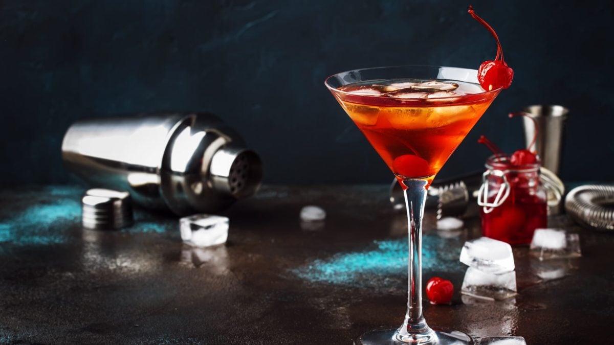 Ein Manhattan-Cocktail in einem Martiniglas, dekoriert mit einer Cocktailkirsche. Im Hintergrund ein umgekippter Cocktail-Shaker und verstreut die Zutaten.