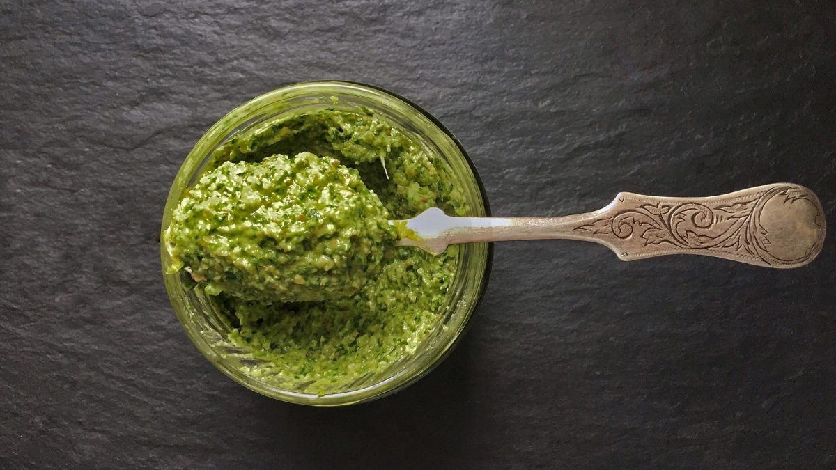 Ein Glas Möhrengrün Pesto mit einem Löffel vor dunklem Hintergrund, von oben.
