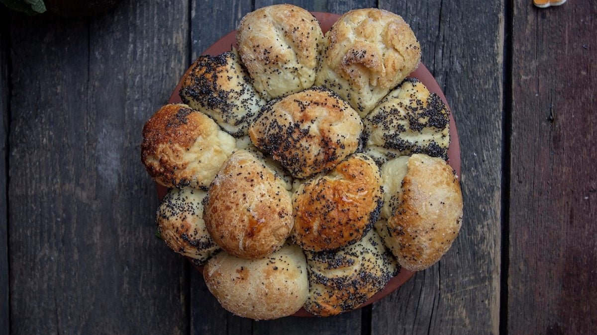 Ein Teller mit einem Monkey-Bread: Kleine Teigkugeln mit Mohn und Salz umhüllt zusammen gebacken. ein einziges großes Brot bildend, vor einem dunklen Holz-Hintergund, von oben.
