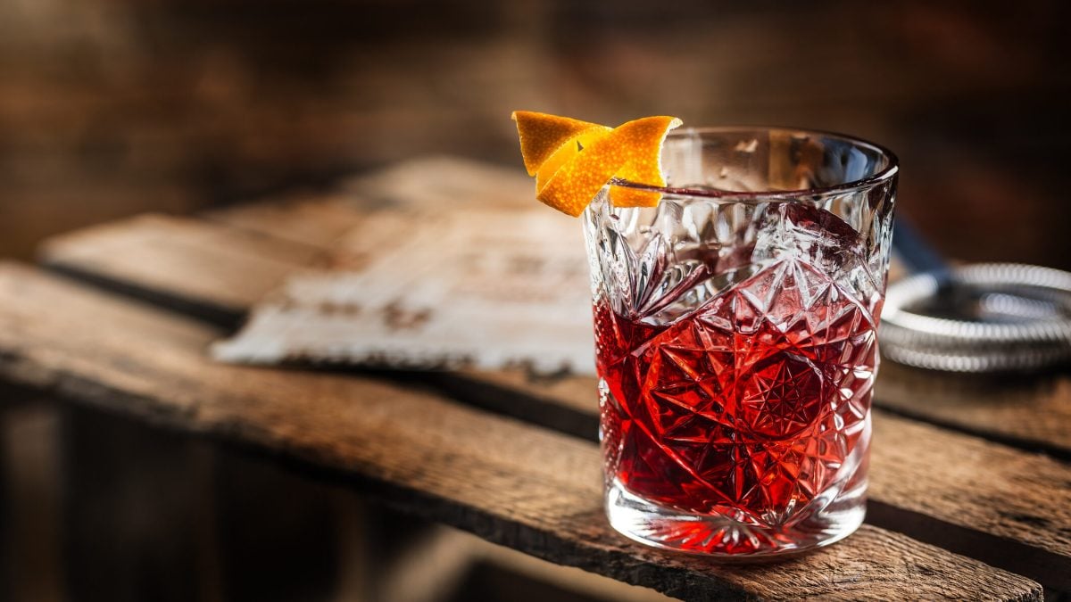 Ein Tumbler-Glas gefüllt mit Negroni und dekoriert mit Orangenschale steht auf einer Weinkiste aus Holz.