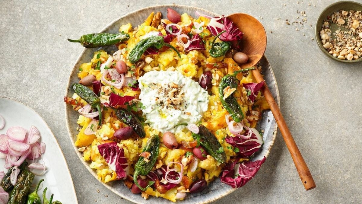 Der orientalische Stampfkartoffelsalat bunt auf einem großen Teller angerichtet. Im Hintergrund stehen Gewürze, Datteln, Jalapenos und Öl.