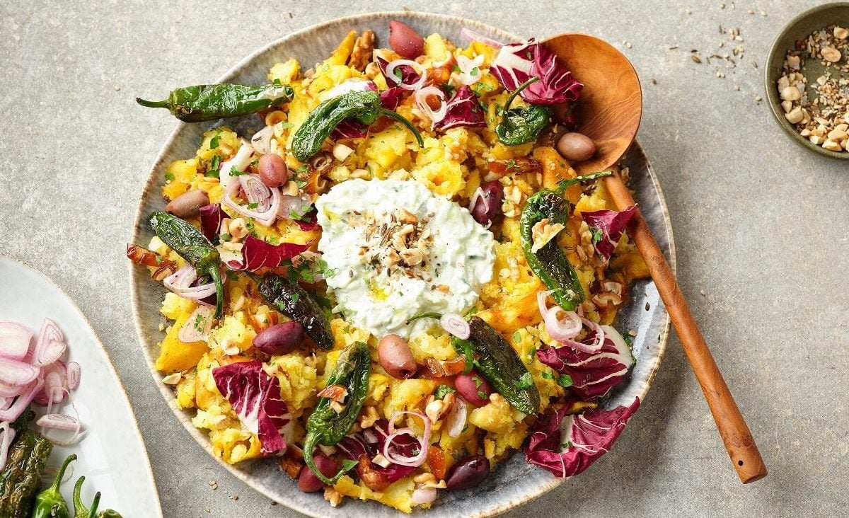 Der orientalische Stampfkartoffelsalat bunt auf einem großen Teller angerichtet. Im Hintergrund stehen Gewürze, Datteln, Jalapenos und Öl.