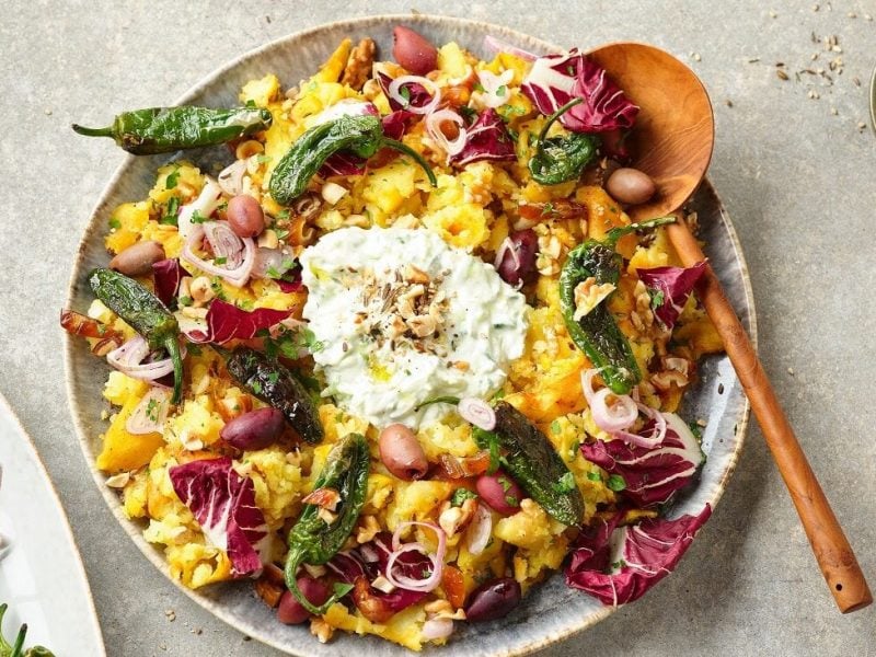 Der orientalische Stampfkartoffelsalat bunt auf einem großen Teller angerichtet. Im Hintergrund stehen Gewürze, Datteln, Jalapenos und Öl.