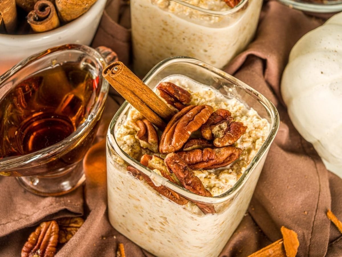 Overnight-Oats mit Ahornsirup und Nüssen auf einem braunen Tuch. Die Oats sind angerichtet mit Pekanüssen und einer Zimtstange. Um die Schale herum stehen ein Zierkürbis, Ahornsirup und Zimtstangen. Auf dem Tuch luegen weitere Pekanüsse.