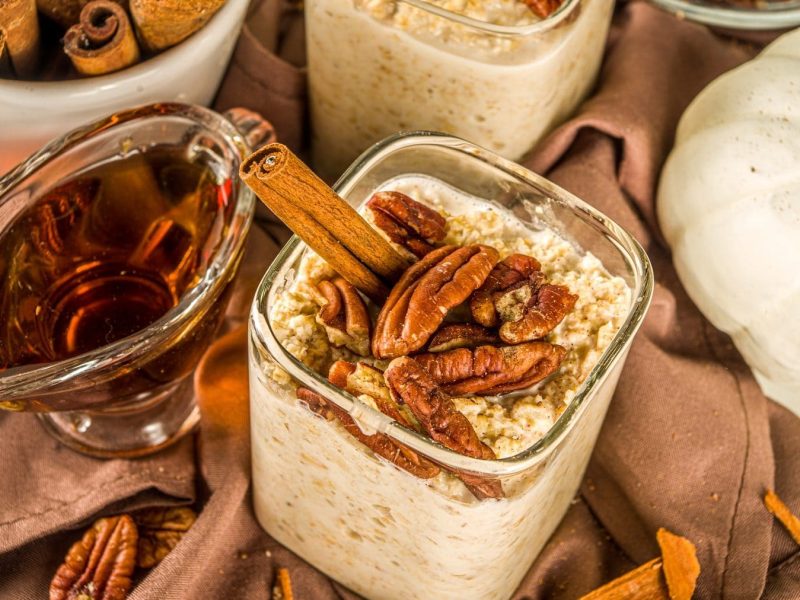 Overnight-Oats mit Ahornsirup und Nüssen auf einem braunen Tuch. Die Oats sind angerichtet mit Pekanüssen und einer Zimtstange. Um die Schale herum stehen ein Zierkürbis, Ahornsirup und Zimtstangen. Auf dem Tuch luegen weitere Pekanüsse.
